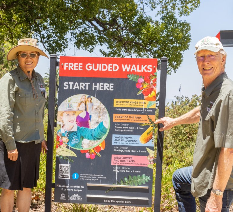 Two volunteer guides smile while standing next to a sign that advertises free guided walks at Kings Park